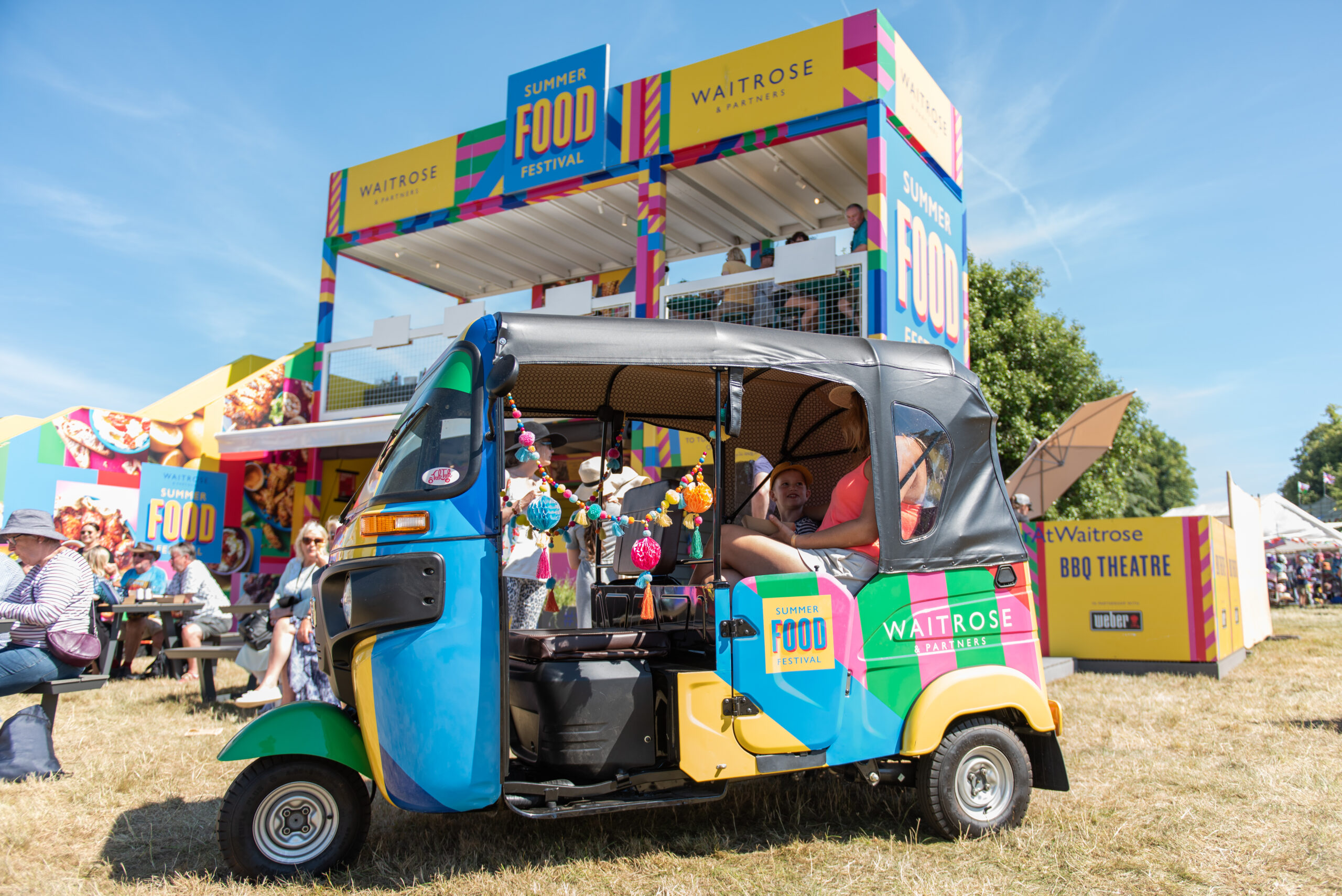 A colorful tuk-tuk decorated with Summer Food Festival logos is parked on grass in front of a vibrant Waitrose booth at the Waitrose Summer Festivals. People are sitting nearby, enjoying the sunny outdoor event.
