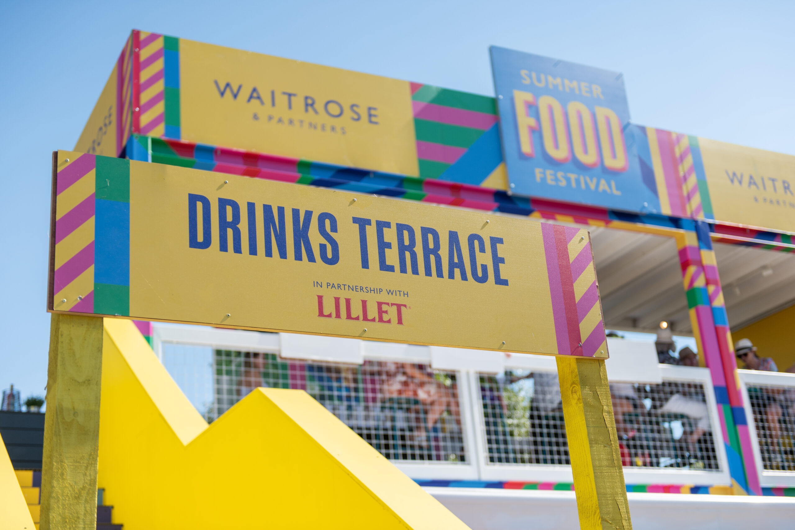 A colorful sign reads Drinks Terrace in partnership with Lillet in front of a vibrant Waitrose Summer Festivals building under a clear blue sky.