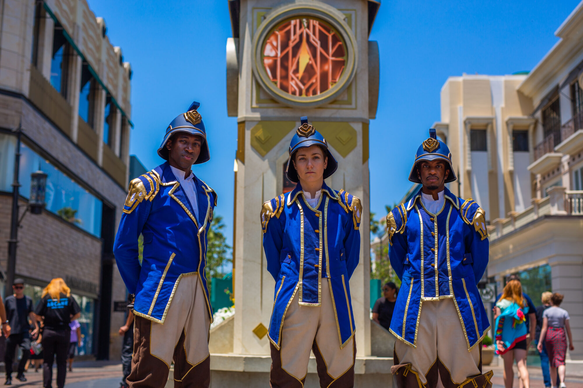 Three people dressed in matching blue and gold fantasy-style guard uniforms, inspired by Arcane FYC Emmys, stand before a decorative clock tower outdoors, with clear blue sky and various people walking by in the background.