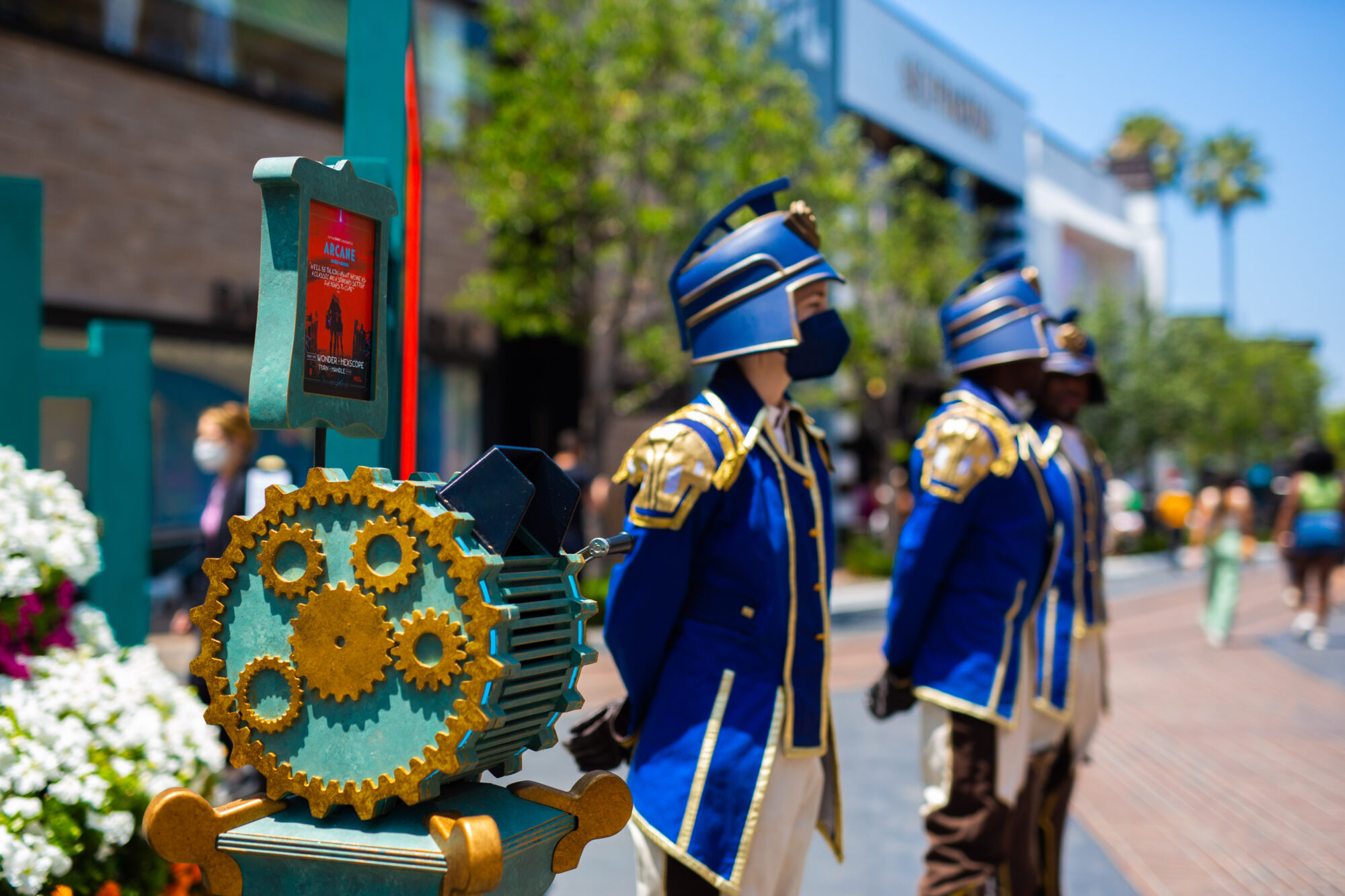 Three people in blue and gold marching band uniforms stand near a machine with colorful gears, as others stroll by in a sunny urban setting—bringing to mind the vibrant, inventive spirit of Arcane FYC Emmys celebrations.