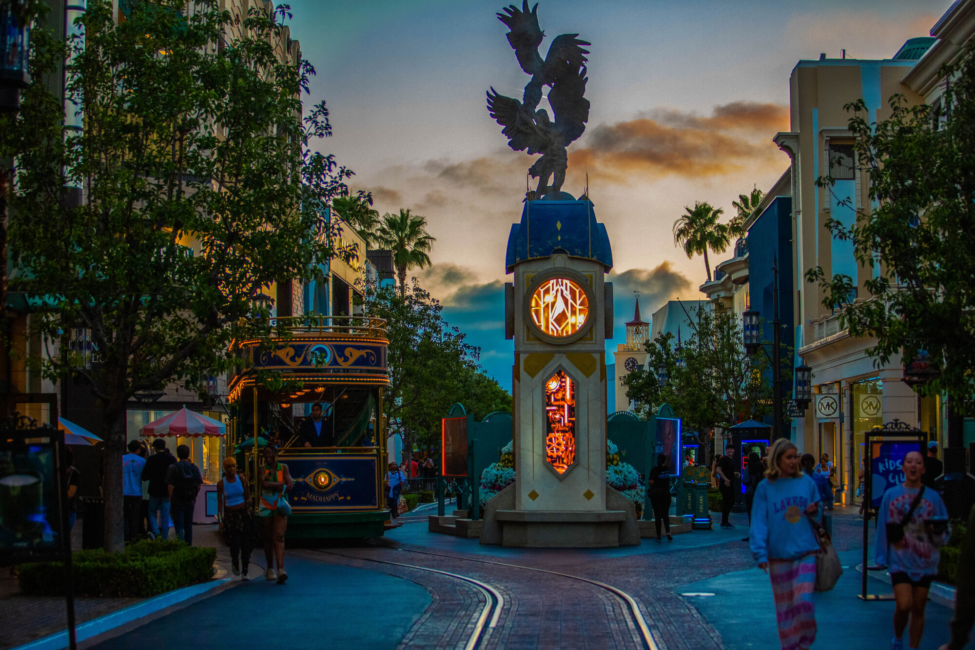 A lively street scene at sunset features a clock tower with a winged statue on top, a vintage trolley, palm trees, and people walking along the tracks amidst storefronts and colorful lights reminiscent of an Arcane FYC Emmys celebration.