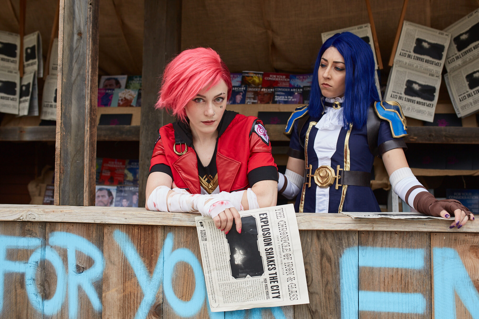 Two people in detailed Arcane cosplay costumes, one with short pink hair and red outfit, the other with long blue hair and blue outfit, stand behind a wooden counter holding newspapers with bold FYC Emmys headlines.