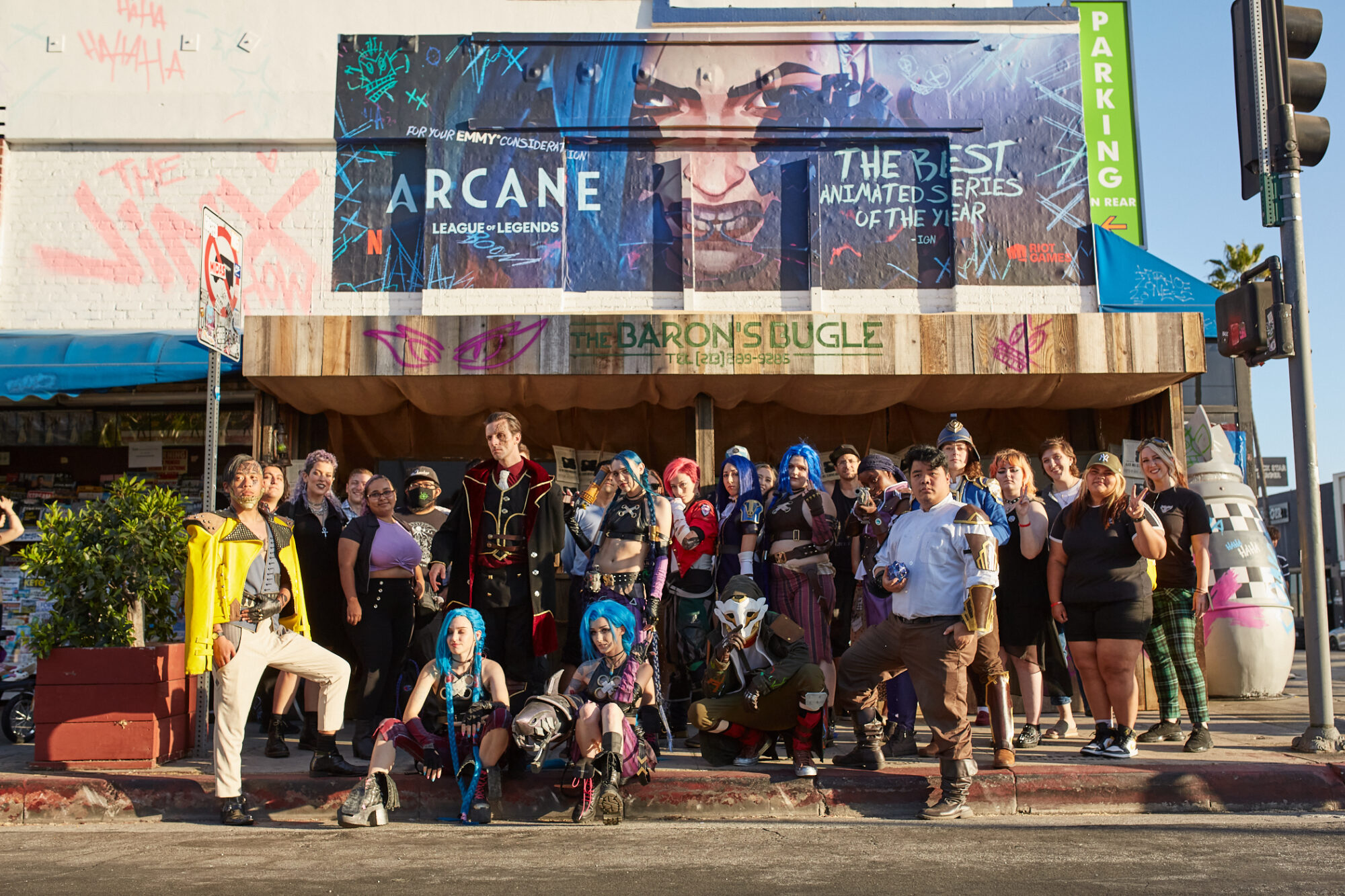 A group of people in detailed Arcane and League of Legends cosplay pose on a city sidewalk in front of a building with a large Arcane FYC Emmys banner above the entrance on a sunny day.