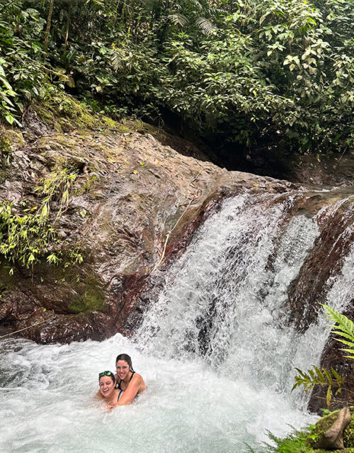 Two people from the marketing team smile and enjoy swimming at the base of a small waterfall, surrounded by lush green tropical plants and rocks.