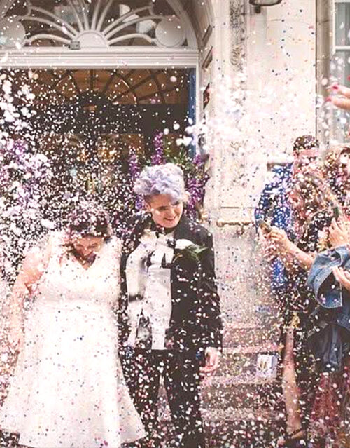 Two newlyweds, one in a white dress and the other in a black suit with short purple hair, smile as they walk outside while their marketing team celebrates and guests throw colorful confetti over them.