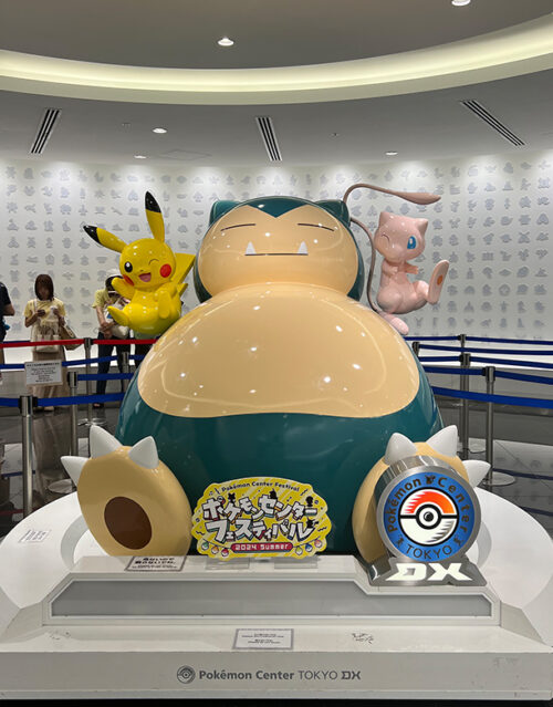 A large Snorlax statue, curated by the marketing team, is displayed at Pokémon Center Tokyo DX, with Pikachu and Mew statues behind it. Signs and a Pokémon Center logo are visible in front, while people stand behind a barrier in the background.