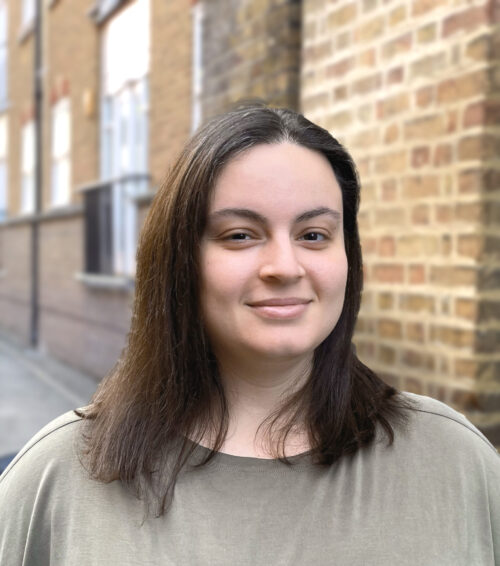 A woman with straight brown hair and a beige top, part of a marketing team, smiles slightly while standing in front of a brick wall and an alleyway.