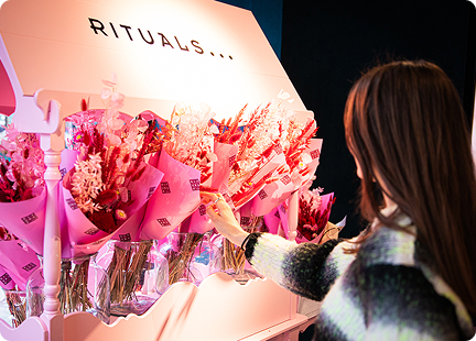 A person with long brown hair selects a pink bouquet from a display stand filled with several pink-wrapped flower arrangements under a sign that says RITUALS, celebrating Rituals Mother’s Day in style.