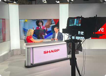 Two people sit at a news desk in a brightly lit TV studio with SHARP on the desk. A camera films them as they discuss Inspire Expo, while a screen behind shows a smiling woman using a laptop.