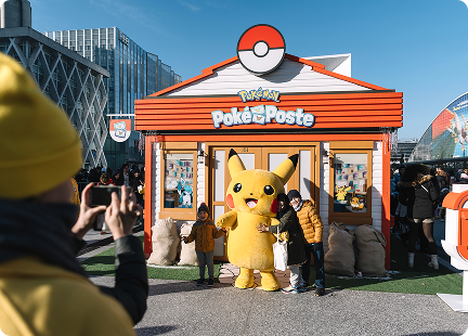 A person snaps a photo of children posing with a Pikachu mascot in front of a Pokémon Poke Post building, surrounded by a lively crowd on a sunny day.
