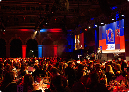 A large, elegant banquet hall filled with seated guests watching a speaker on stage at the Top Employer Awards. The room is illuminated with red and blue lights, and a big screen displays event graphics behind the stage.