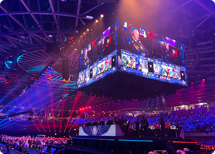 A large esports arena filled with spectators features a central, hanging screen displaying a Red Bull LOIO video game competition. Colorful lights and stage setups create an energetic atmosphere, with players seated below the main screen.