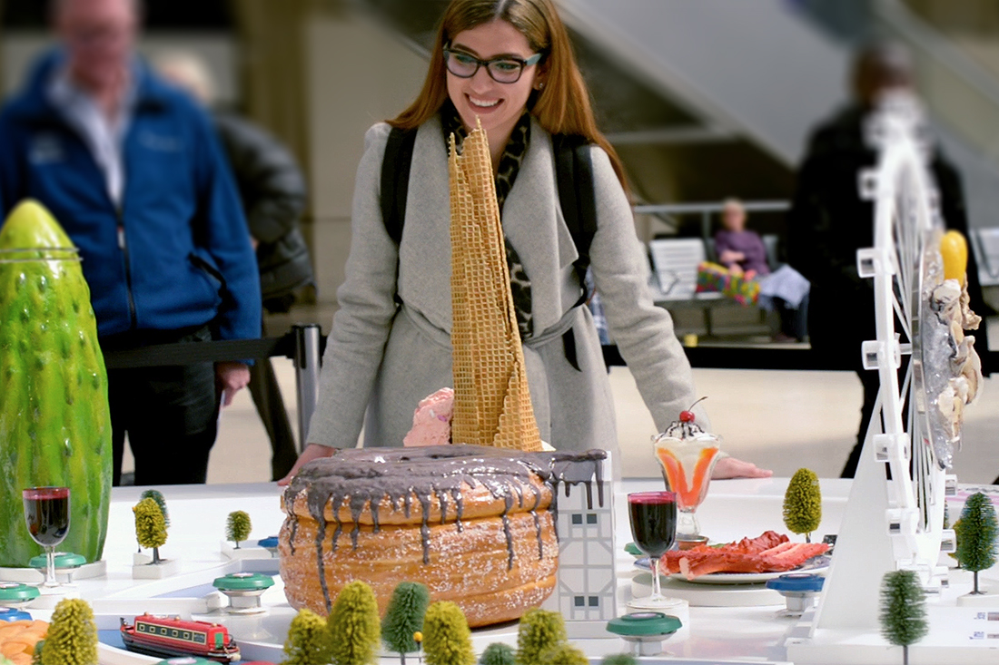A woman in glasses and a gray coat smiles at a whimsical Bookatable case study display featuring giant pastries, desserts, wine glasses, and miniature models of famous landmarks, trees, and a small red bus.