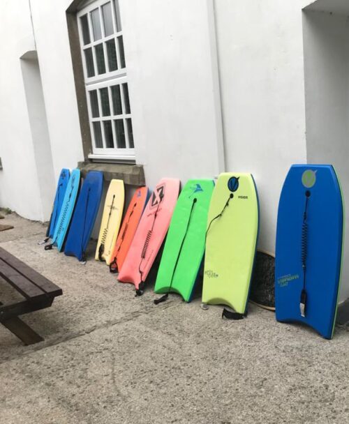 Nine colorful bodyboards are lined up against a white wall outside, next to a wooden picnic table and a large window—like supplies ready for a marketing team’s creative beach outing. The boards come in blue, orange, pink, green, and yellow.
