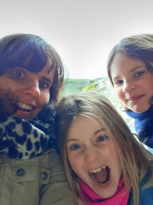 A woman and two young girls, looking like a close-knit marketing team, pose for a selfie outdoors. The woman and one girl smile at the camera, while the other girl makes a playful, wide-mouthed expression in their cozy winter clothes.