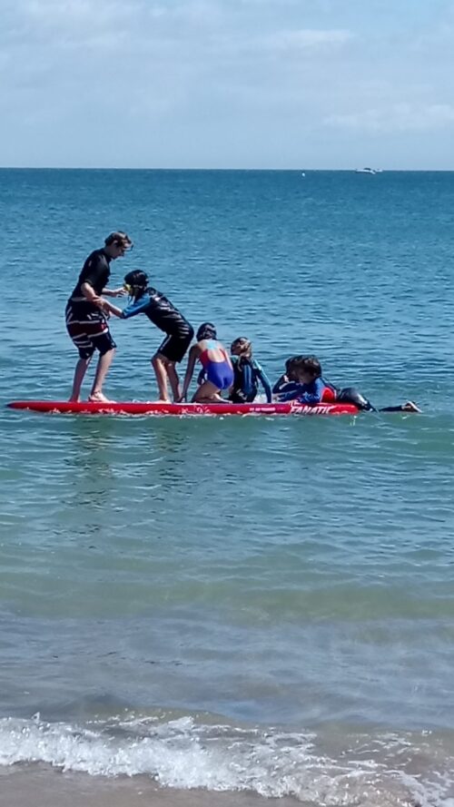 Six children, resembling a dynamic marketing team, are on a large red paddleboard in the ocean. Some are sitting, some kneeling, two standing, and one climbs back on. The sky above is partly cloudy near the shore.