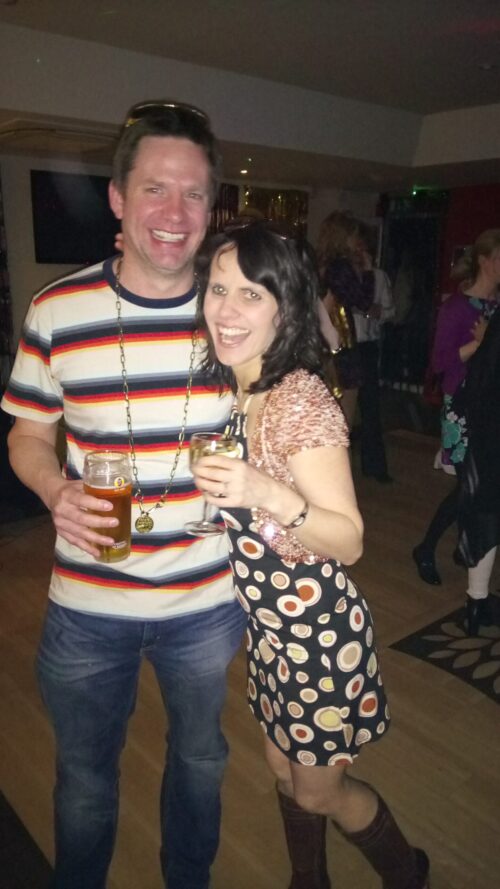 A man and woman from the marketing team smile and pose together at a party. The man holds a pint of beer, wearing a striped shirt and necklace; the woman wears a patterned dress with brown boots, holding wine. Others mingle in the background.