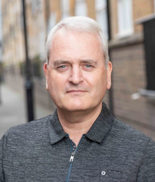 A middle-aged man with short gray hair, part of a marketing team, is wearing a dark zip-up sweater and standing outdoors in front of a blurred, urban brick street background.