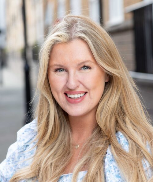 A woman with long blonde hair and light blue eyes smiles outdoors. Wearing a light blue patterned top and a small gold necklace, she stands on a blurred city street, looking confident—perfect for representing your marketing team.