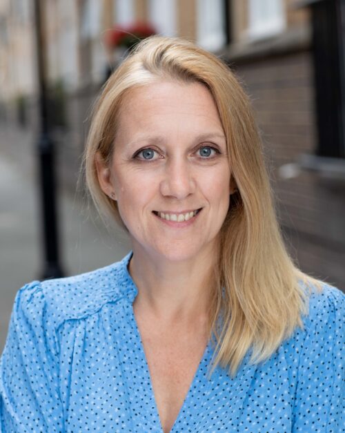 A smiling woman with straight blonde hair, part of the marketing team, wears a light blue blouse with small polka dots as she stands outdoors, with a blurred brick wall and street in the background.