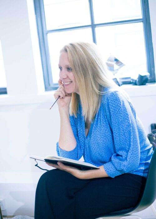 A woman with long blonde hair, wearing a blue blouse and black pants, sits by a window, smiling and holding a notebook and pen, appearing deep in thought while brainstorming ideas for her marketing team.