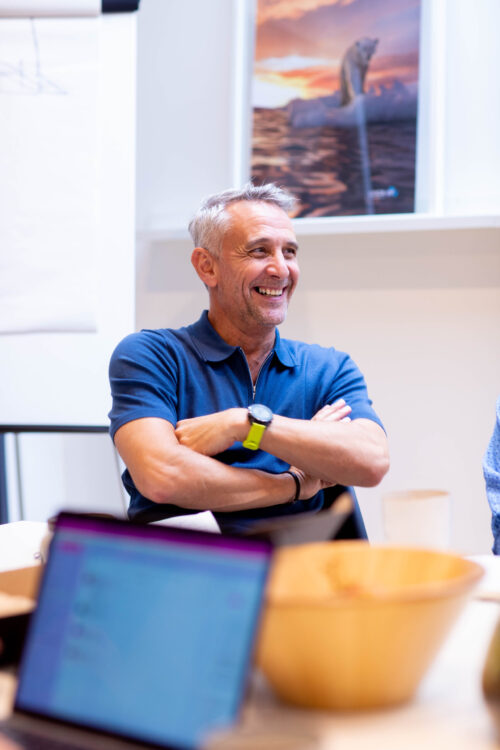 A smiling man with gray hair sits at a table with his arms crossed, wearing a blue polo shirt and green watch. A laptop hints at his work with the marketing team, while a wooden bowl and polar bear poster add character to the scene.