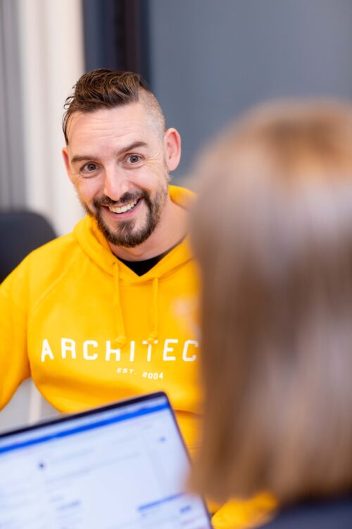 A smiling man in a yellow hoodie that reads ARCHITECT talks to another person from the marketing team, who is blurred in the foreground and using a laptop.