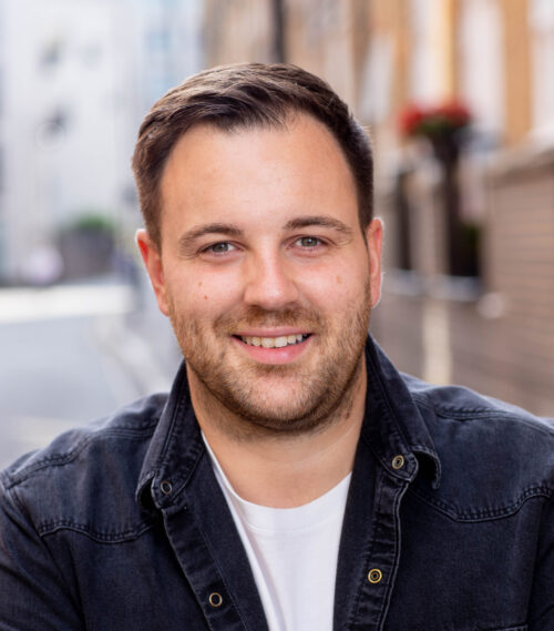 A man with short brown hair and a trimmed beard smiles at the camera, representing the marketing team. He is wearing a black denim jacket over a white shirt, with a blurred urban street scene in the background.