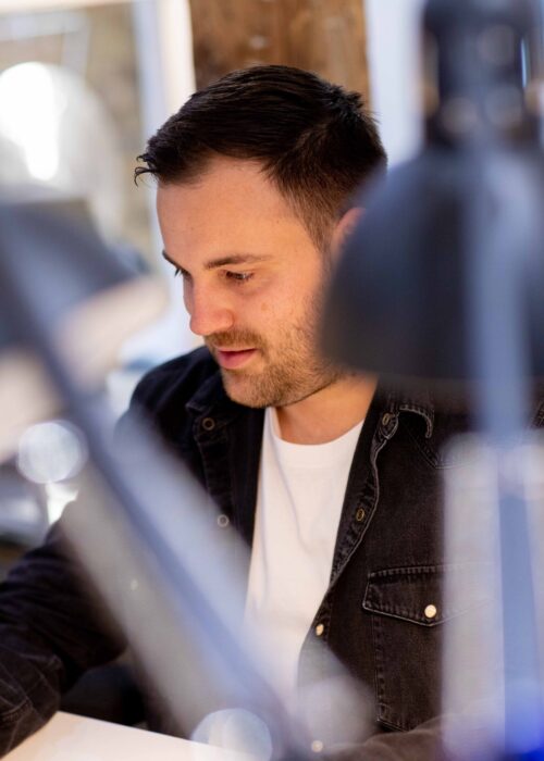 A man with short dark hair and a beard, wearing a dark shirt over a white t-shirt, sits at a desk and focuses on his work with the marketing team. The foreground features blurred desk lamps.