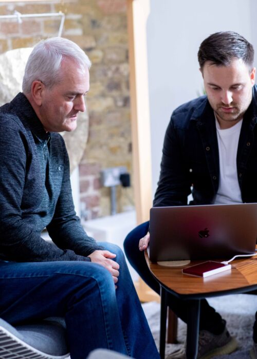 Two men sit together indoors, focused on a laptop atop a small wooden table. The older and younger men appear to be part of a marketing team, engaged in collaborative discussion or work.