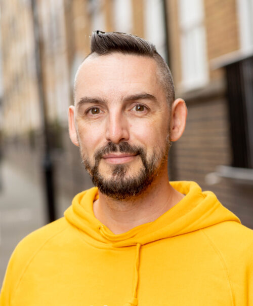 A man with short dark hair, a beard, and mustache, wearing a bright yellow hoodie, stands outside on a city street with blurred brick buildings in the background. He is smiling slightly and looking at the camera, as if ready to join the marketing team.