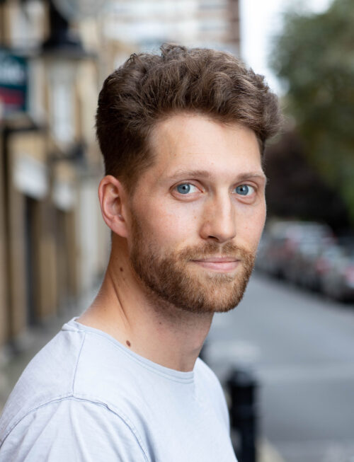 A man with short curly brown hair, a beard, and blue eyes stands outdoors on a city street in a light gray shirt, looking at the camera with a neutral expression—an approachable member of the marketing team.