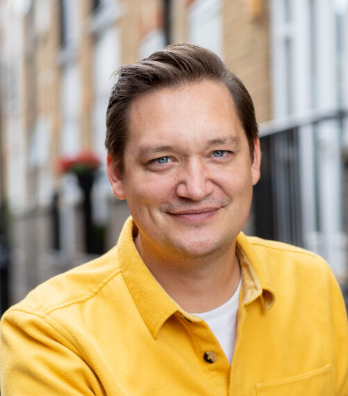 A man with light brown hair and blue eyes, part of the marketing team, smiles at the camera. He is wearing a yellow jacket over a white shirt and is outdoors, with blurred buildings and railings in the background.