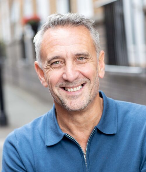 A middle-aged man with short gray hair and a beard, part of the marketing team, smiles warmly at the camera while wearing a blue zip-up shirt. The background is an outdoor urban street with blurred buildings and railings.