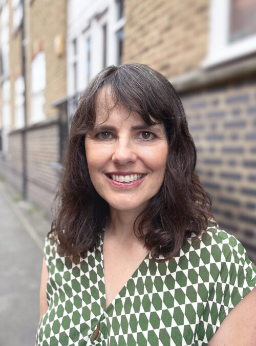 A woman with wavy brown hair and a patterned green and white top smiles at the camera. She is standing outdoors in front of a brick building, representing her marketing team with confidence and warmth.