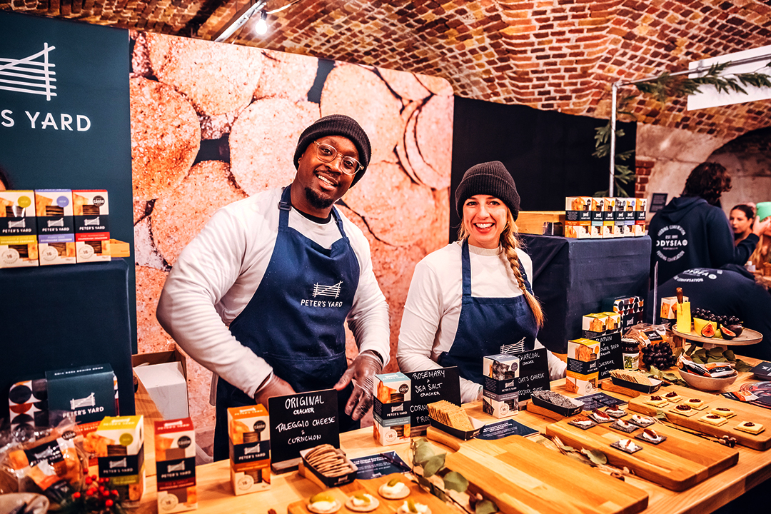 Two smiling vendors wearing navy aprons and black beanies stand behind a display of packaged snacks and samples at a Waitrose Winter Festival food market stall with a brick wall background.
