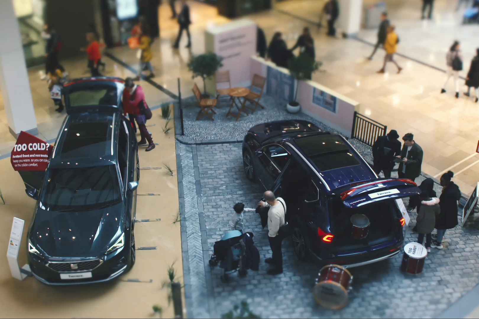 A top-down view of a SEAT case study car exhibition in a mall, with two cars on display, people interacting, and tables and chairs set up nearby. Shoppers walk around in the background.