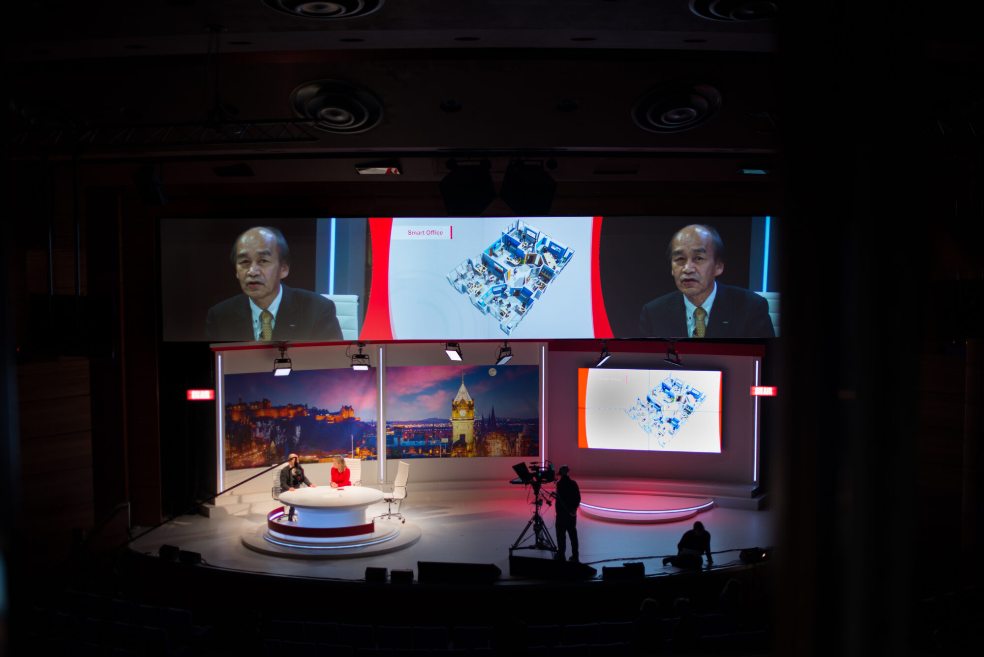 A stage set for Inspire Expo features two people seated at a round table and a host standing nearby, while large screens above display close-up shots and presentation slides. A camera crew films the scene.