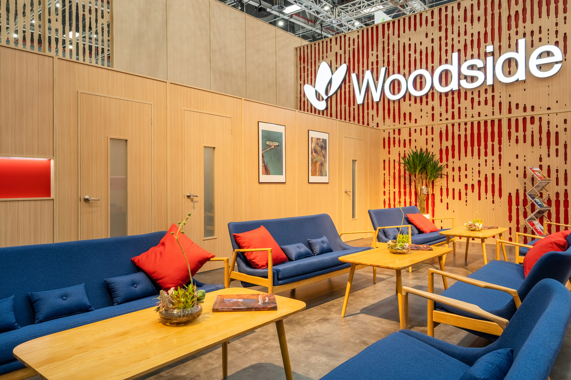 Modern lounge area with blue sofas, red and orange cushions, wooden tables, potted plants, and framed artwork. The wall features the Woodside logo and a red decorative pattern on light wood paneling for a standout Woodside case study.