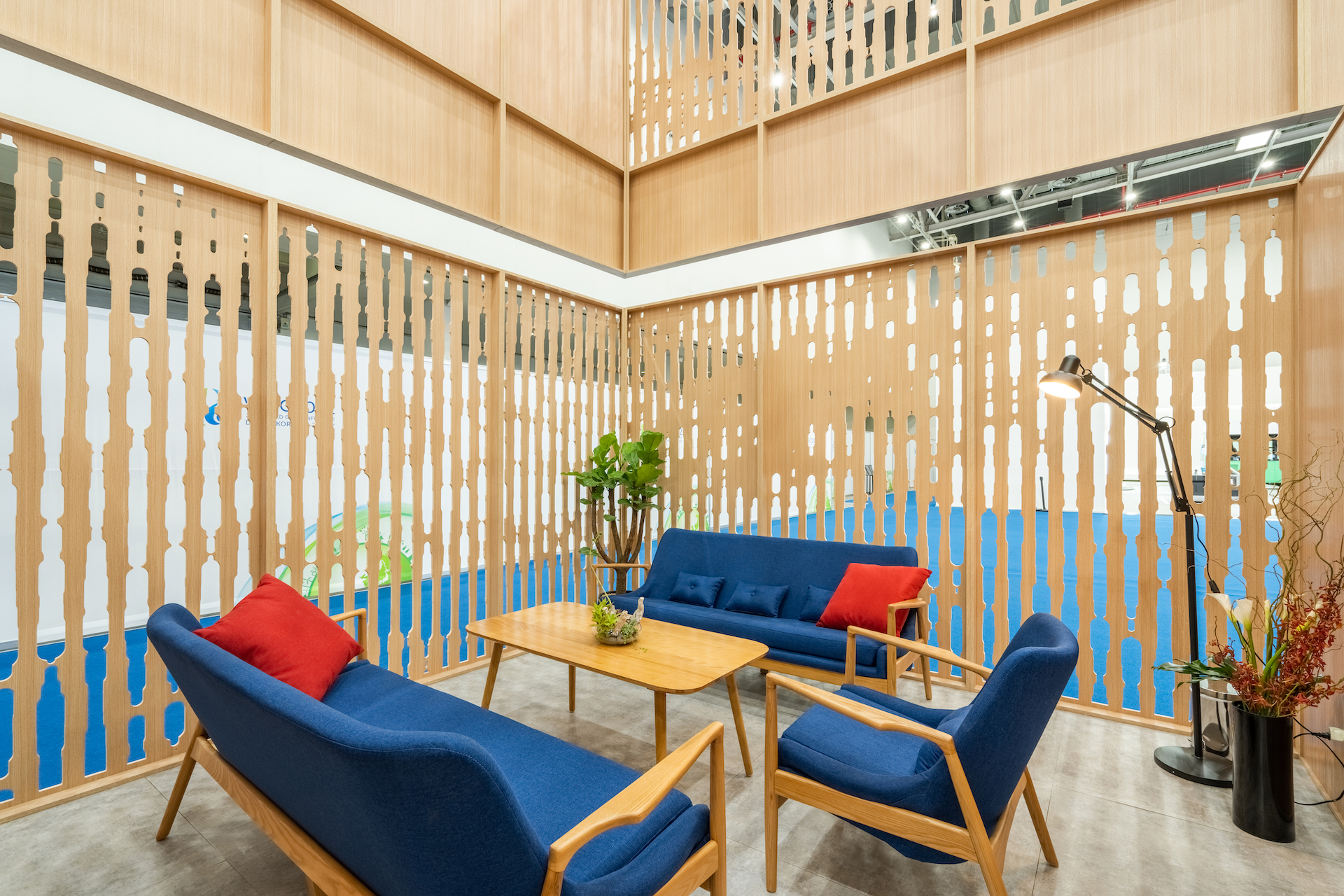 A modern lounge area with blue sofas and wooden furniture, accented by red and orange throw pillows. Inspired by the Woodside case study, decorative wooden slats enclose the space, while plants and a floor lamp add a cozy touch.