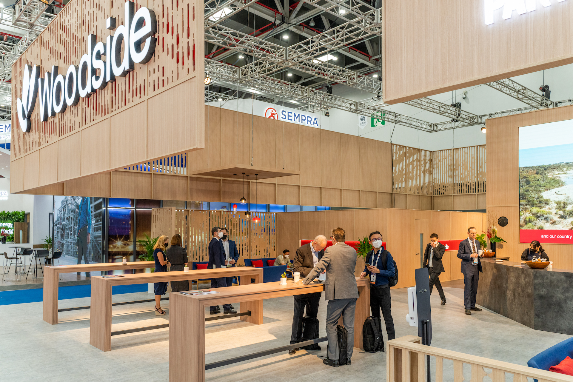 A modern exhibition booth with a large Woodside sign showcases a Woodside case study, featuring wooden tables and people in business attire networking and talking in a bright, open convention hall.