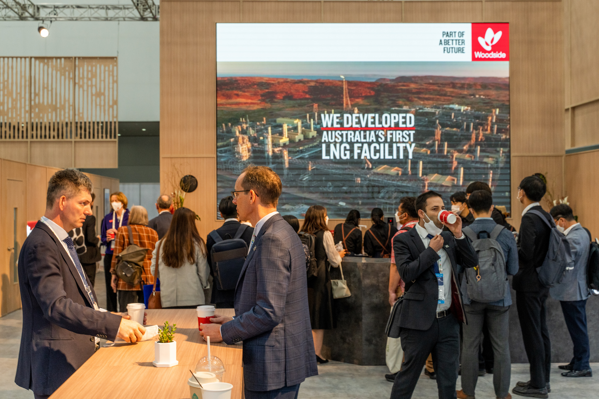 A group of people in business attire interact at an indoor event. Behind them, a large screen displays “We developed Australia’s first LNG facility” with the Woodside logo—a scene from the Woodside case study. Some people are holding drinks and networking.