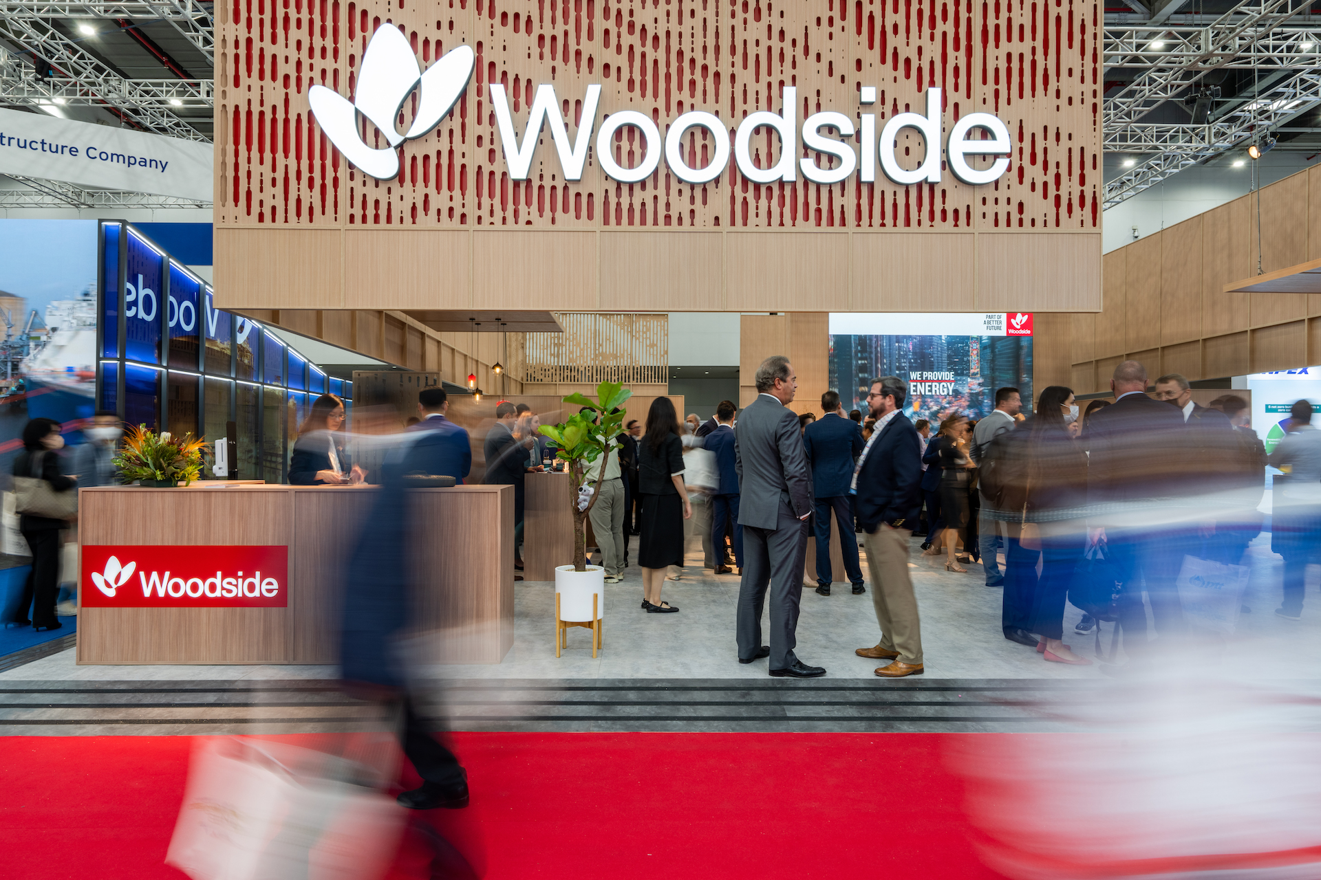 People attend a busy exhibition booth for Woodside, featuring a large logo and modern wooden design. Attendees interact and walk past, highlighting the success captured in this Woodside case study, with some motion blur on people in the foreground.