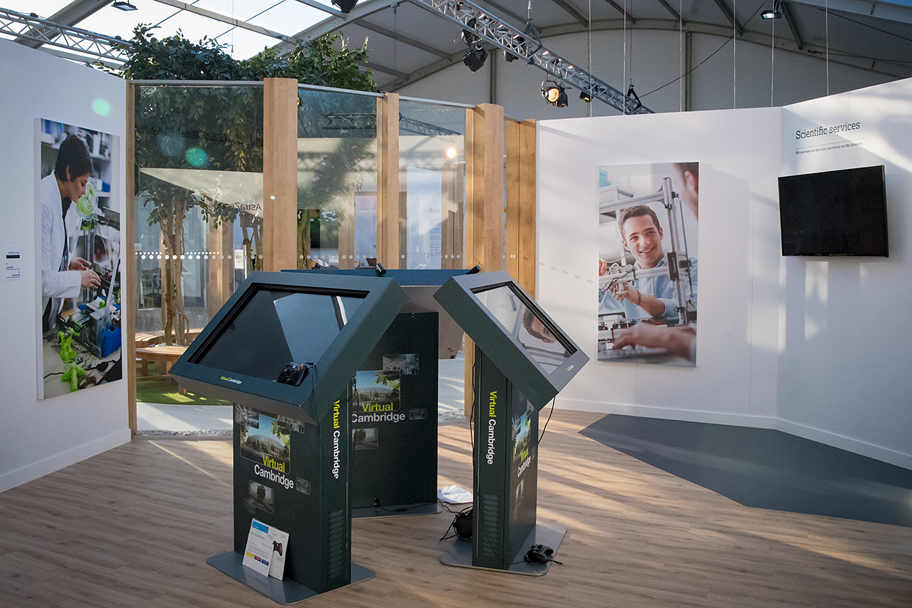 A modern exhibition booth highlights the AstraZeneca RNG partnership with two interactive touchscreen kiosks, large wall photos of smiling scientists in laboratories, wood accents, and a TV mounted on a white wall.