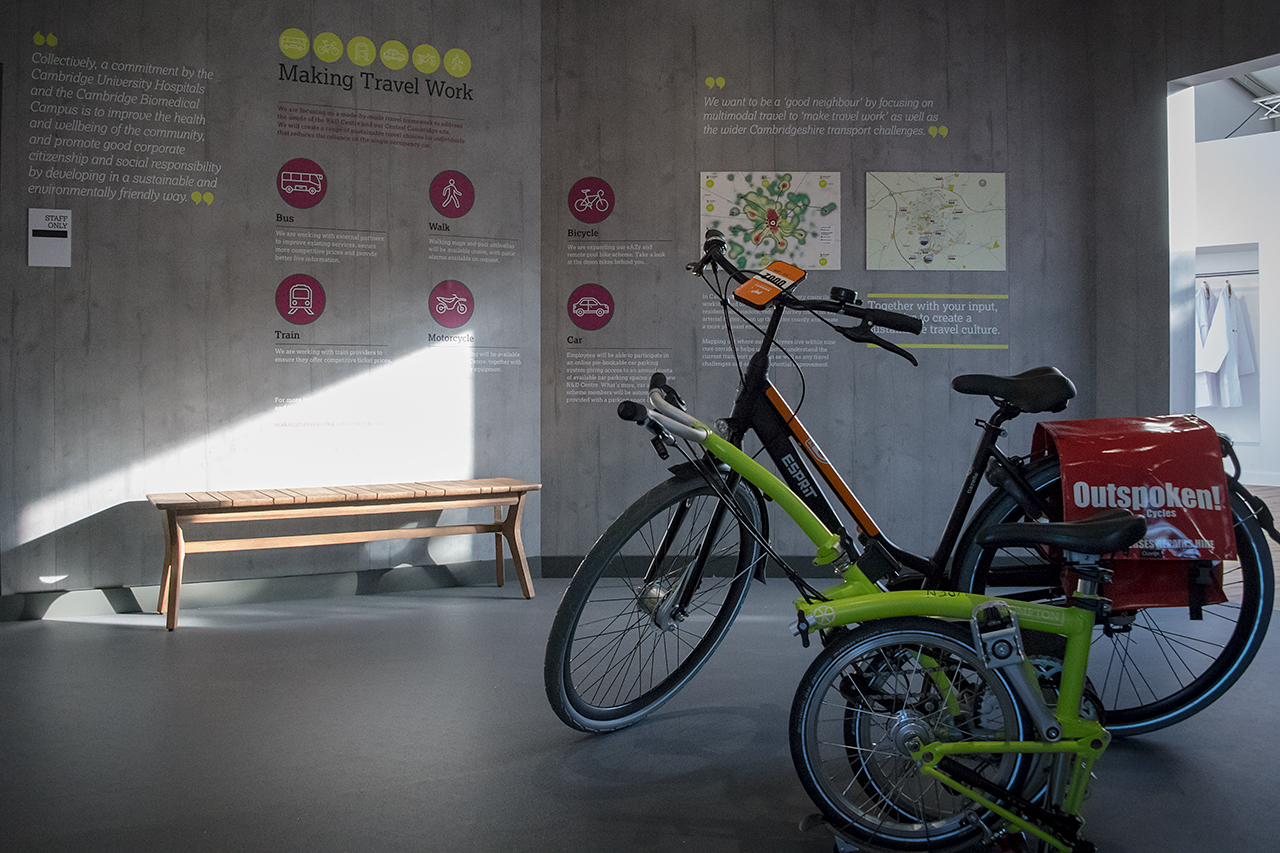 Two bicycles, one with an Outspoken! delivery bag, stand in a well-lit indoor space. Behind them is a wooden bench and a wall display featuring sustainable travel options and the AstraZeneca RNG partnership for greener commuting.
