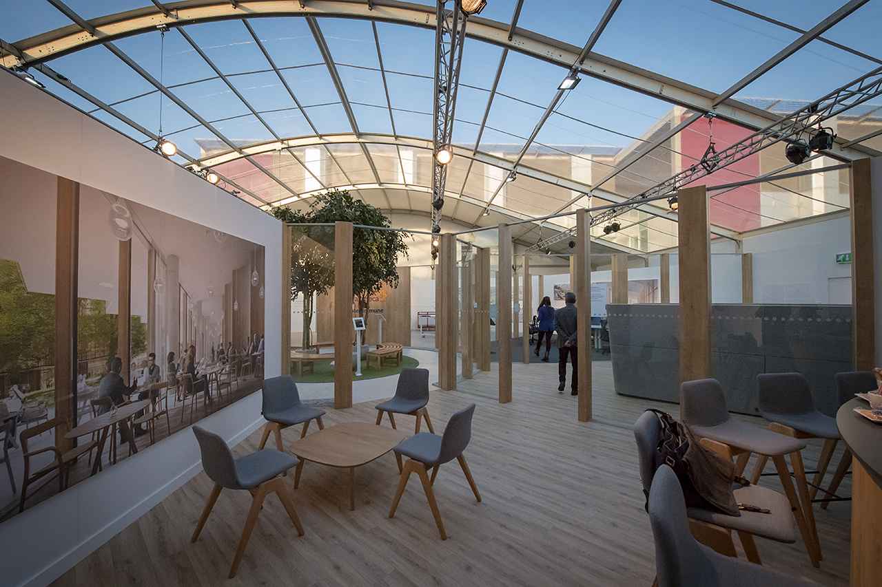 Spacious modern indoor area with wooden floors, gray chairs around small tables, large wall art, and glass walls under a curved transparent roof—ideal for meetings like the AstraZeneca RNG partnership. A few people are seen in the background.