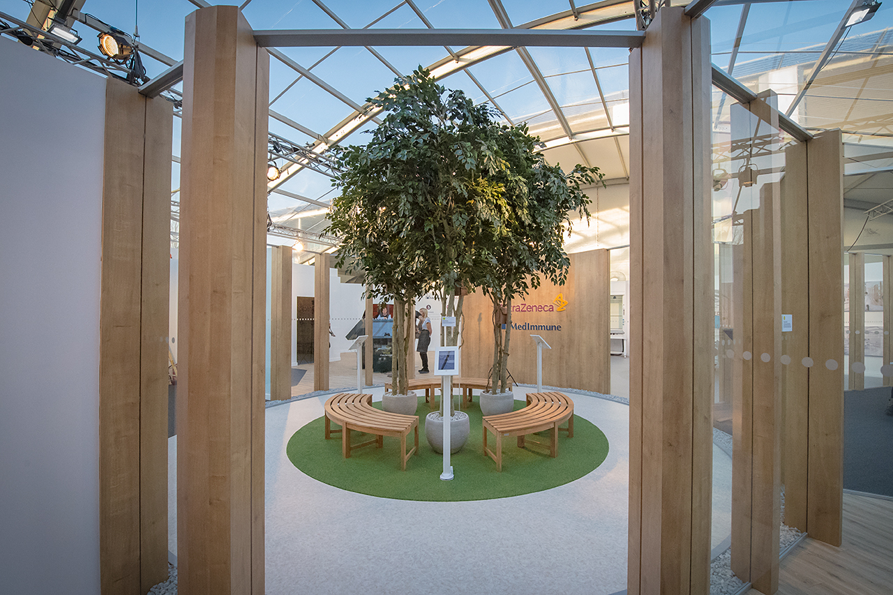 A modern indoor space with a large potted tree in the center, featuring curved wooden benches on a circular green carpet—an inviting area enclosed by glass walls and wooden beams, inspired by the AstraZeneca RNG partnership’s commitment to sustainability.