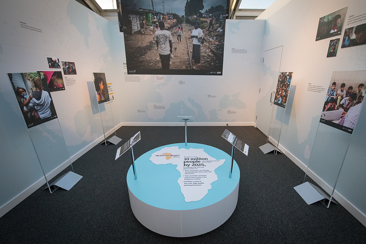 Museum exhibit room with photos and stands, featuring a central platform with a map of Africa and text about malnourished children, highlighting hunger awareness by 2025 and the AstraZeneca RNG partnerships role in driving positive change.