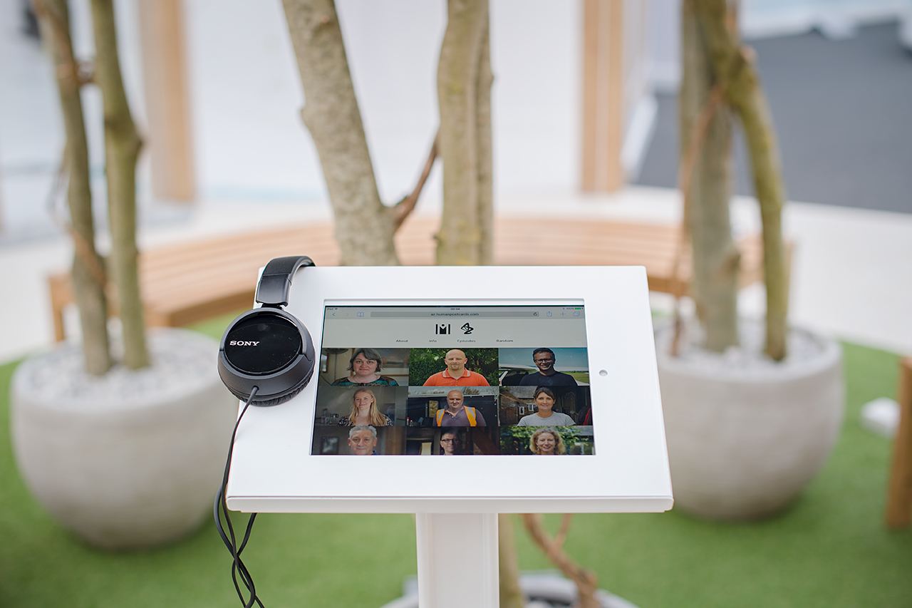 A tablet on a white stand displays a video call interface, possibly discussing the AstraZeneca RNG partnership, with Sony headphones resting on it. In the background, potted trees and curved wooden benches sit on green artificial grass.
