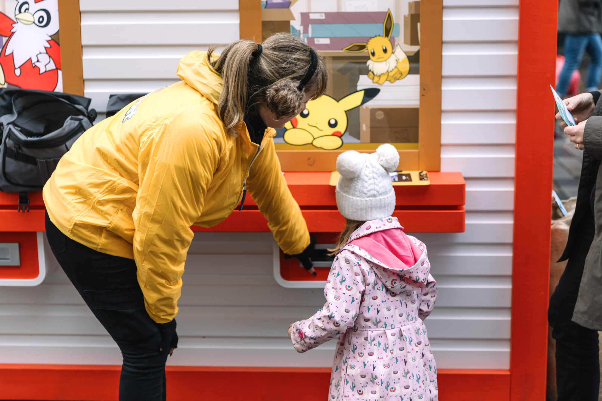 A woman in a yellow jacket helps a young child in a white hat and pink coat at a Pokémon Poke Post kiosk decorated with images of Pikachu and Eevee.